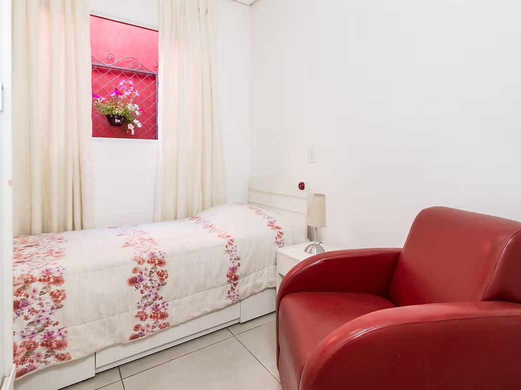 Quarto individual do Residencial Espaço Saúde com cama de colcha florida, poltrona vermelha, abajur e cortinas claras
