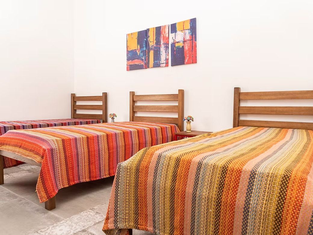 Quarto da La Vie com três camas de madeira, colchas listradas em tons de laranja e vermelho e um quadro colorido na parede