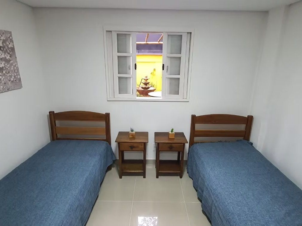 Quarto duplo da Kûara com duas camas de colcha azul, criados-mudos de madeira e uma janela ao centro