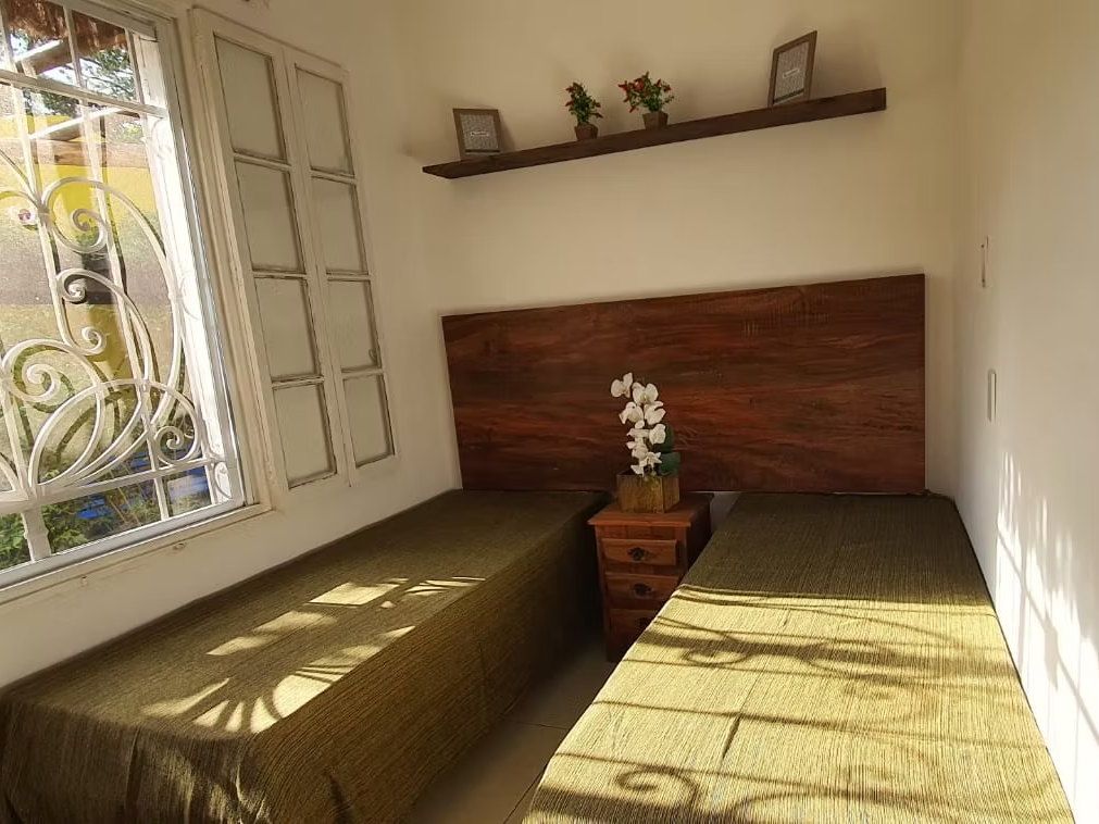 Quarto duplo da Kûara com cabeceira de madeira, orquídea no criado-mudo e sol entrando pela janela de grade rendada