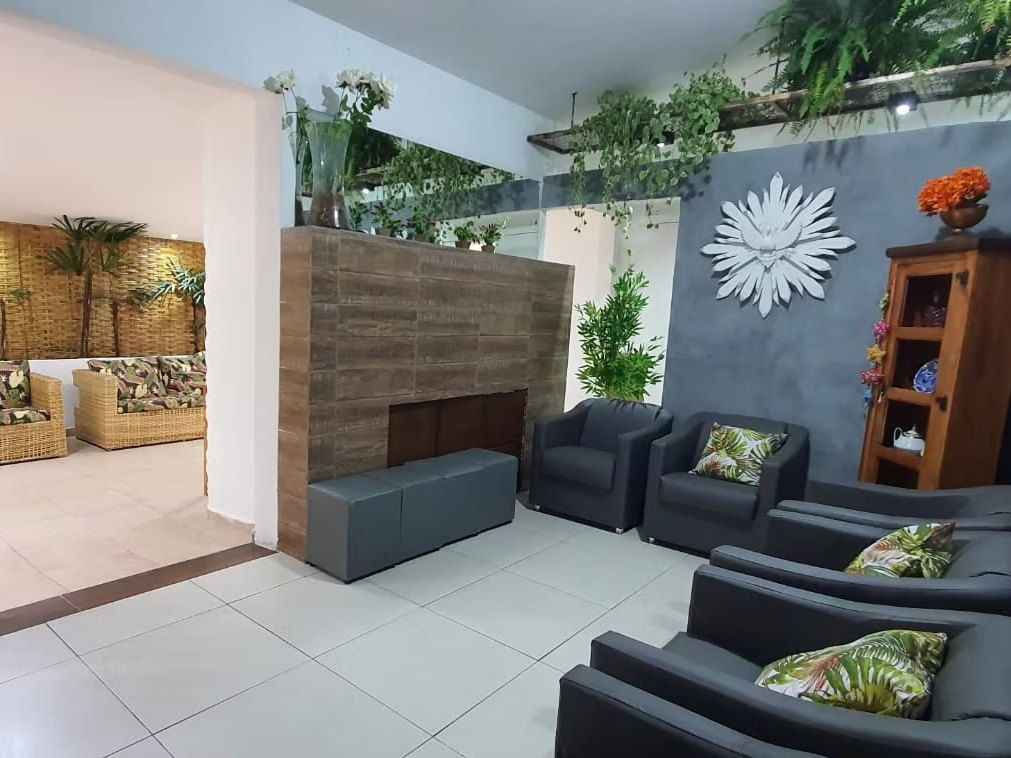 Sala de convivência da Kûara com poltronas cinza e almofadas de folhagem, parede cinza com espelho em forma de sol e estante de madeira