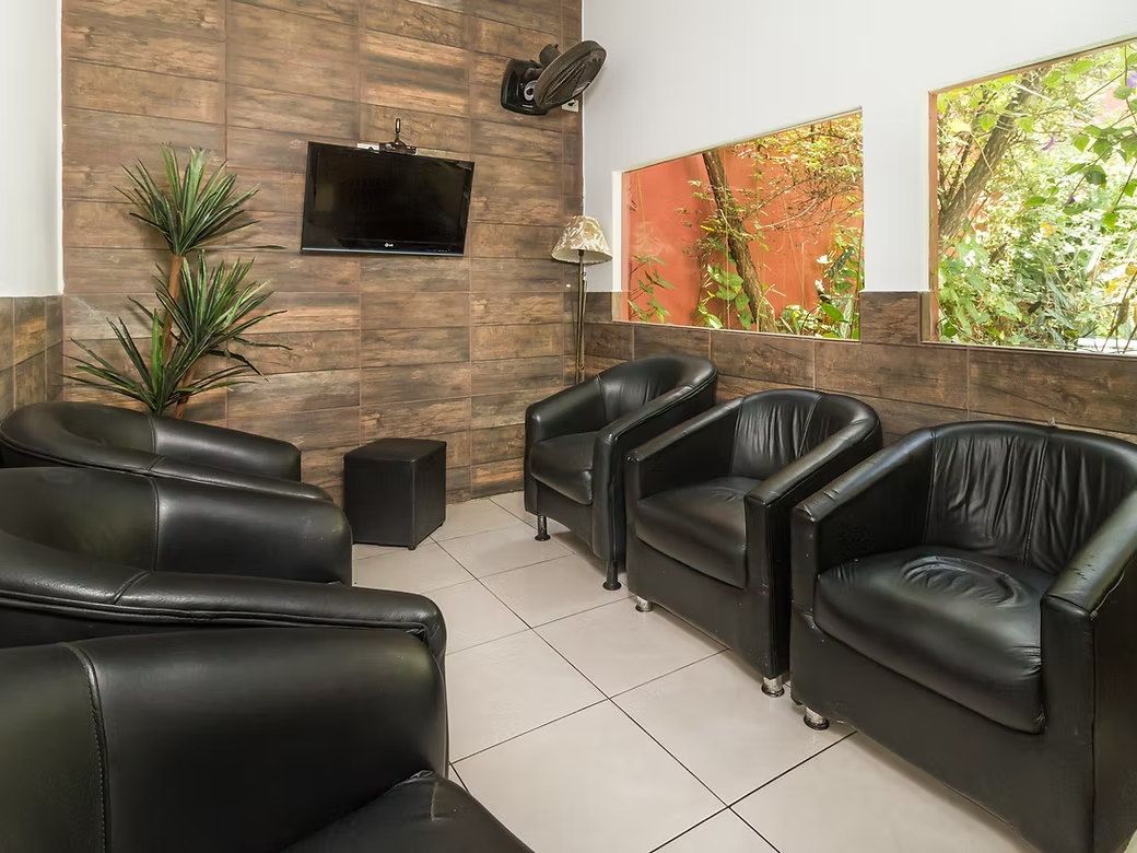Sala de convivência da Tsuru com poltronas pretas, TV na parede de madeira e janelas abertas para o jardim